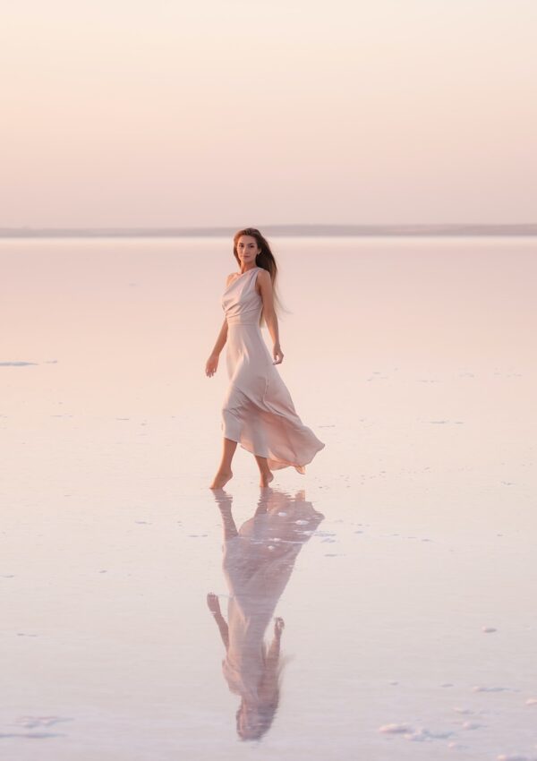 Young blonde woman in evening pastel pink, powdery dress stands barefoot on white crystallized salt